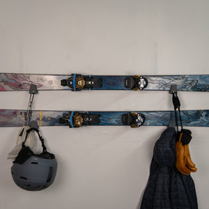 Skis with bindings, helmet, gloves, and jacket hanging on a white wall.