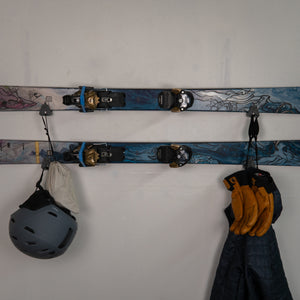 Skis with bindings, helmet, and gloves hanging on a white wall.