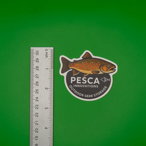 Brown trout and 'PESCA Innovations' sticker on a green background, accompanied by a ruler for scale, 2.5 inches tall