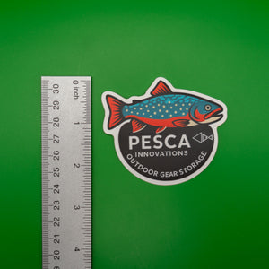 Brook trout and 'PESCA Innovations' sticker on a green background, accompanied by a ruler for scale, 2.5 inches tall