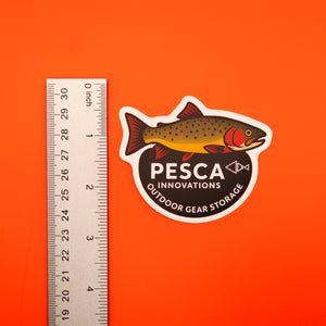 Cutthroat trout and 'PESCA Innovations' sticker on an orange background, accompanied by a ruler for scale, 3 inches tall