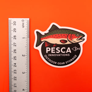 Rainbow trout and 'PESCA Innovations' sticker on an orange background, accompanied by a ruler for scale, 3 inches tall