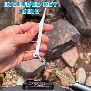 A hand holds a white nail knot tool with key ring with a metal split ring and a coiled yellow-green spring, with rocks in the background and the text 'INCLUDES KEY RING' at the top.